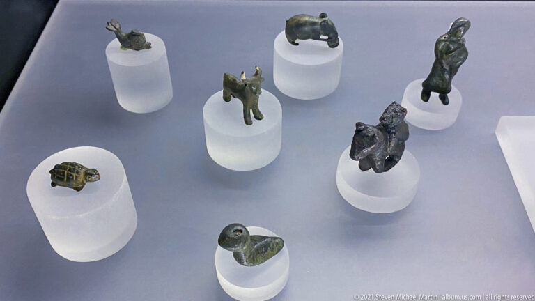 Small bronze animal figurines, including several quadrupeds, arranged on separate white cylindrical mounts in a display case.