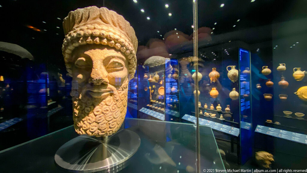 Large bearded male head with elaborate headdress displayed in front of rows of smaller antiquities in a museum case.