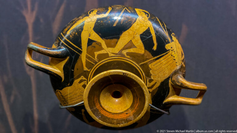 Underside of a red-figure drinking cup with nude male figures around the foot and two horizontal handles.