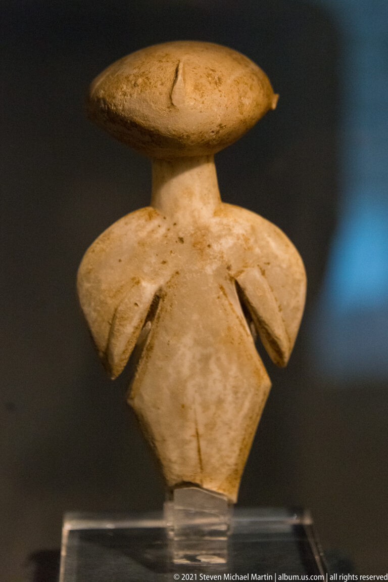 Small pale marble Cycladic figure fragment with oval head, broad shoulders, bent arms, and a tapering broken torso.