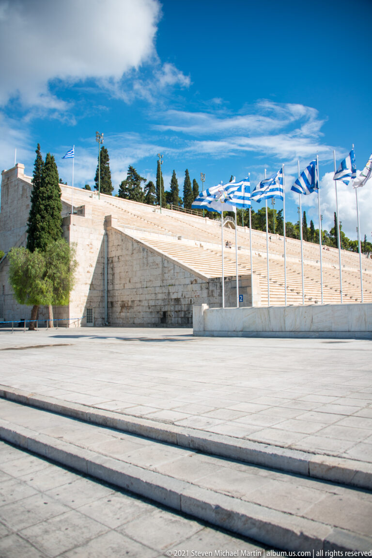 Marble seating tiers of the Panathenaic Stadium behind a paved forecourt lined with Greek flags.