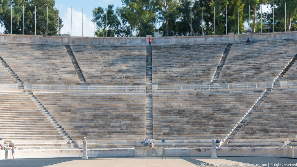 Curved marble grandstand with dark radial stair aisles and a few visitors at the Panathenaic Stadium.
