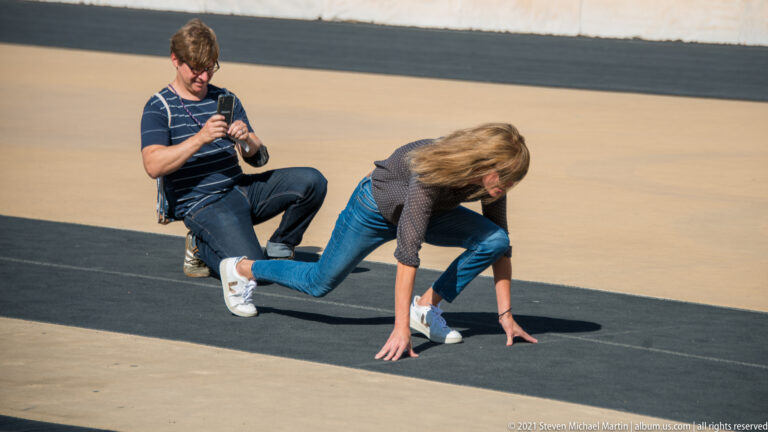 Visitor crouching in a sprint-start pose on the stadium track while another person kneels behind with a camera.