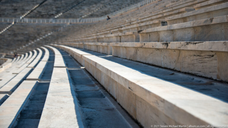 Oblique view along curving marble stadium benches, with alternating bands of sunlight and shadow.
