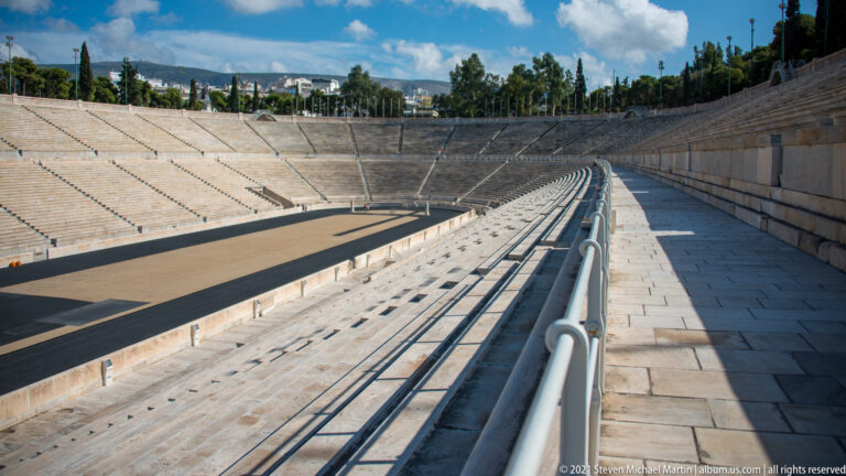 Elevated view along marble seating and handrails toward the curved end of the Panathenaic Stadium and track below.
