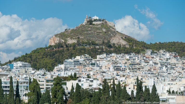 Telephoto view of rocky Mount Lycabettus with summit buildings above densely packed white buildings in Athens.