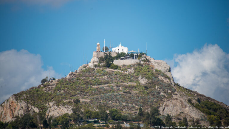 Rocky summit of Mount Lycabettus with white hilltop structures against a blue sky and clouds.
