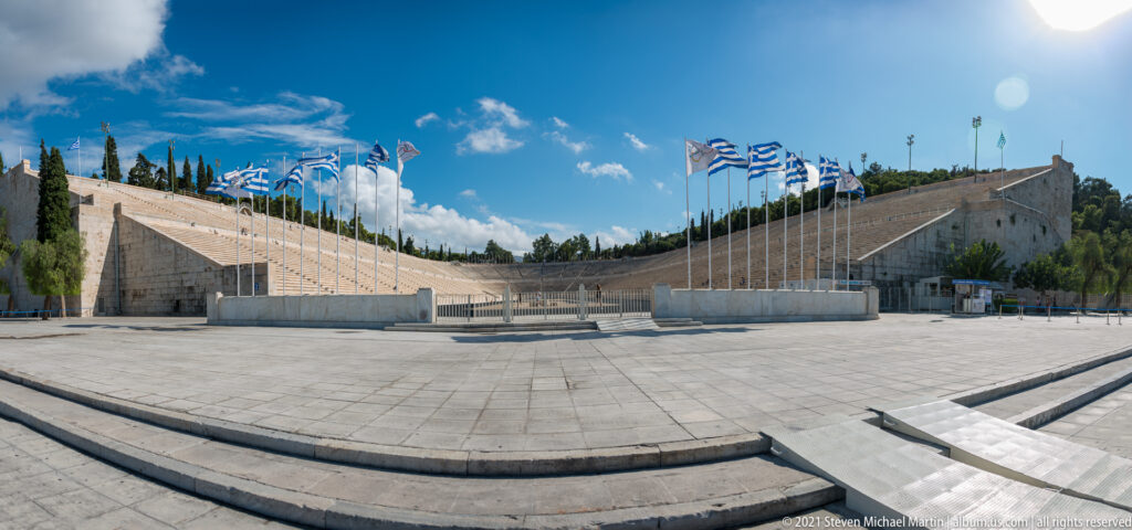 Wide view across the Panathenaic Stadium forecourt toward steep marble stands and rows of Greek and Olympic flags.