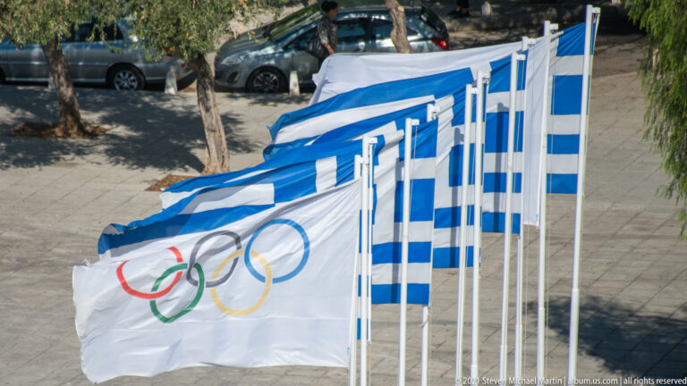 White Olympic flag with colored rings flying among Greek flags, with a street and trees below.