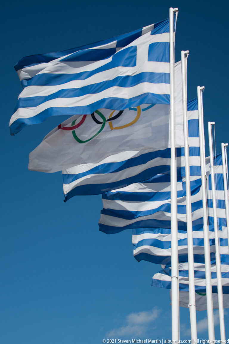 Windblown Greek flags and a partially visible Olympic flag on white poles against a deep blue sky.