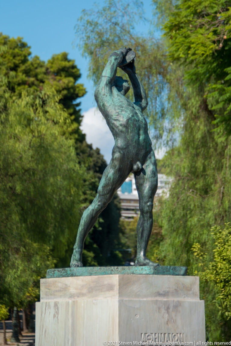 Bronze statue of a nude discus thrower twisting with the discus raised overhead on a stone pedestal among trees.