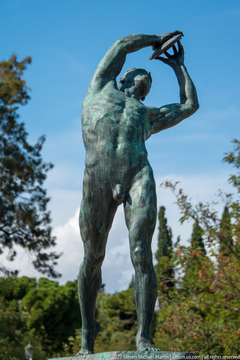 Close low-angle view of a green-patinated bronze discus thrower holding the discus high above his head.