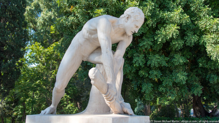 White marble statue of a nude male athlete bending forward with one hand near his lower leg, backed by trees.