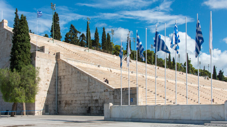 Greek flags on tall poles in front of steep marble seating and a large stair structure at the Panathenaic Stadium.