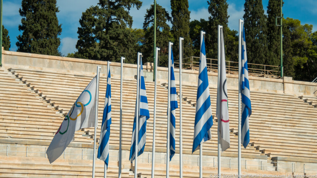 Greek and white Olympic flags on poles against the marble seating tiers of the Panathenaic Stadium.