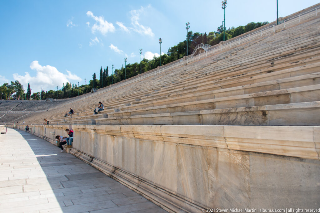 Low-angle view along long marble seating tiers at the Panathenaic Stadium, with a few visitors scattered across the stands.