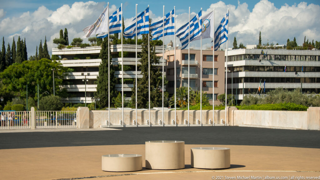Greek flags beside the dark stadium track, with low cylindrical forms, trees, and apartment buildings beyond.
