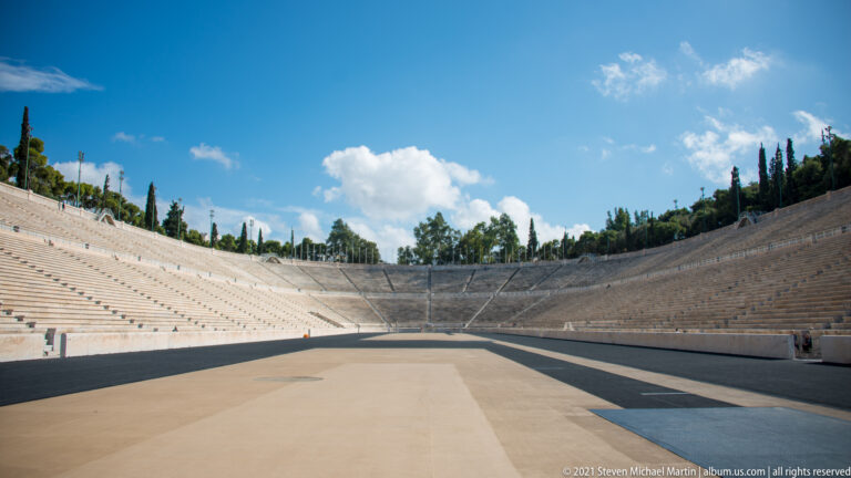 Empty marble seating curves around the closed end of the Panathenaic Stadium beyond tan and black track surfaces.