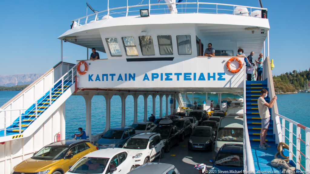 Open-deck Greek car ferry loaded with cars, with passengers on stairs and bridge deck above the vehicle deck.