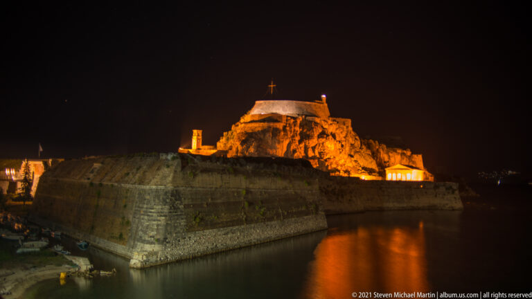 Large stone bastion beside dark water, with the floodlit Old Fortress of Corfu rising behind it at night.