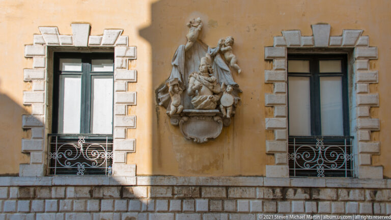 Ocher façade with two tall windows, pale rusticated surrounds, wrought-iron grilles, and a central sculptural relief.