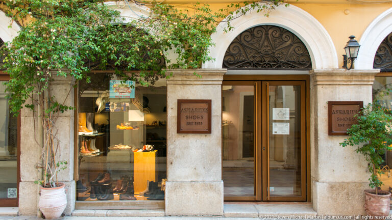 Corfu shoe shop with arched stone frontage, footwear display, glazed door, ornamental fanlights, and potted plants.