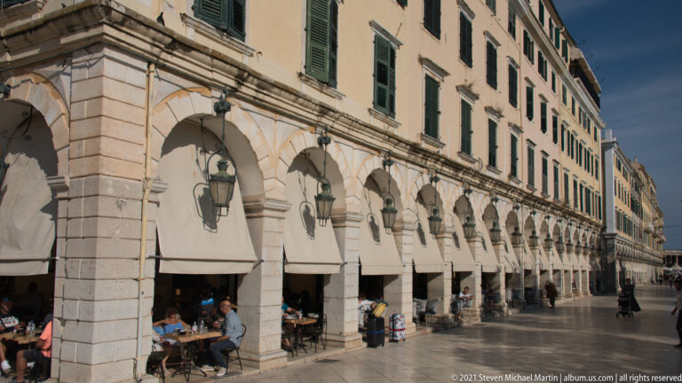 Long Corfu arcade with repeated stone arches, hanging lanterns, café tables, pedestrians, and upper stories with green shutters.