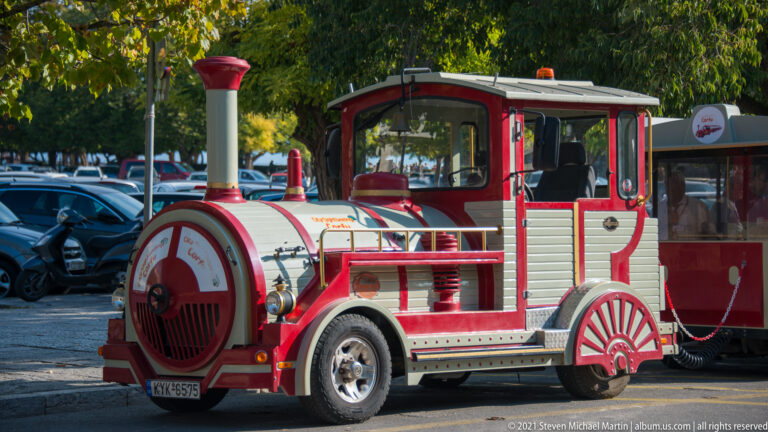 Red-and-cream road train styled like a steam locomotive, coupled to a passenger carriage beside parked cars.