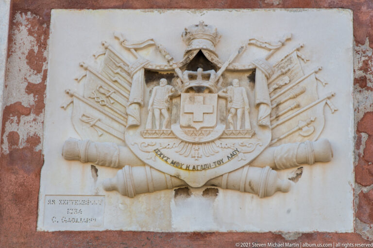 Pale stone armorial relief on red plaster with crowned shield, figures, banners, weapons, drums, and crossed cannon barrels.