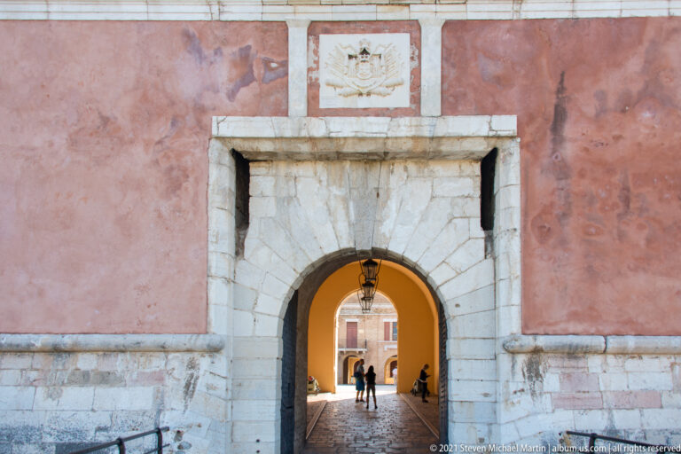 Large arched gateway in a red fortress wall with pale stone surround, armorial relief above, and visitors in the passage.