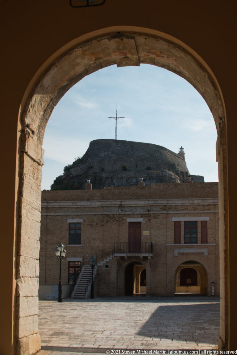 Fortress courtyard building and rocky summit seen through a large shadowed stone arch at Corfu's Old Fortress.