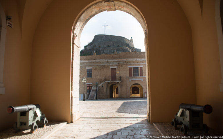 Two cannons inside a dark fortress passage framing a sunlit courtyard building and rocky summit through an arch.