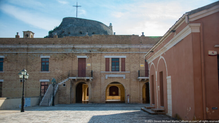 Stone courtyard building at Corfu's Old Fortress with exterior stairs, arched ground-floor openings, and rocky summit behind.