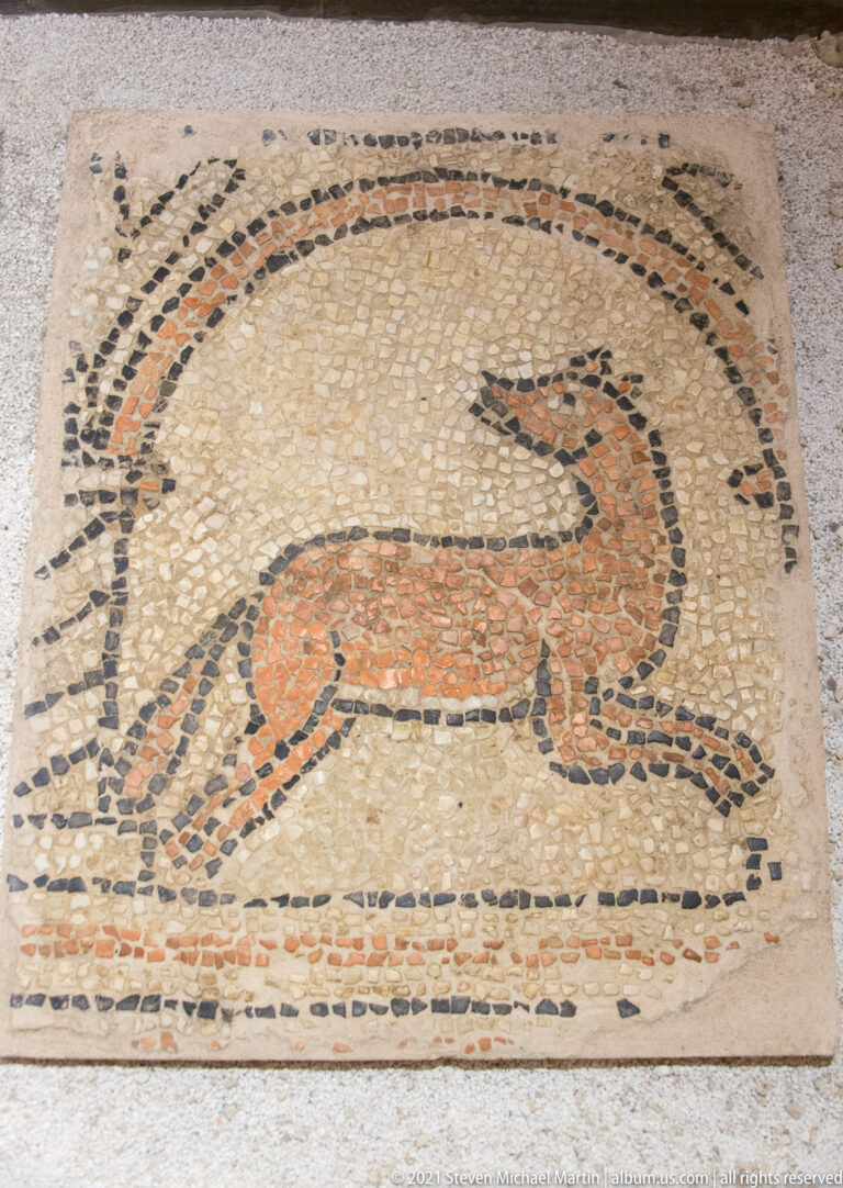 Ancient-style mosaic panel of a running quadruped in black, red, ocher, and cream tesserae within a bordered field.