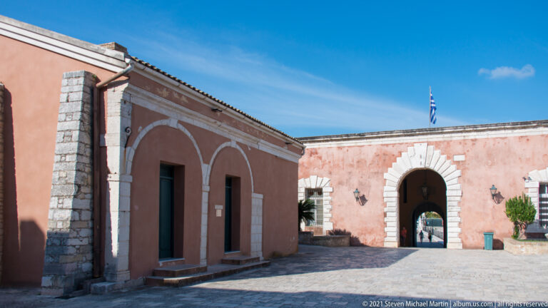 Sunlit Old Fortress courtyard with red-plastered buildings, pale stone trim, arched recesses, gateway, and Greek flag.