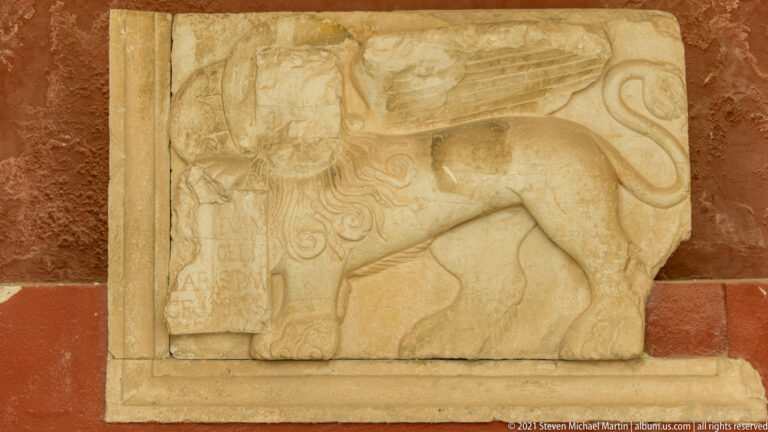 Weathered stone relief of a winged lion beside an open book, mounted against a red wall.