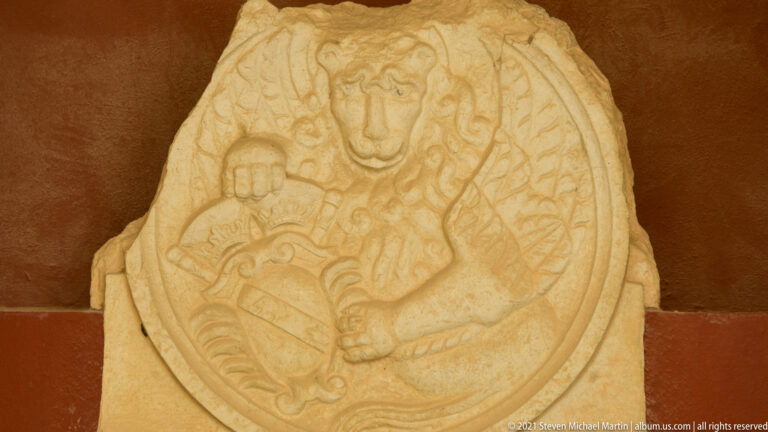 Oval weathered stone relief with a frontal figure, broad wing-like forms, and a rounded shield-like element.
