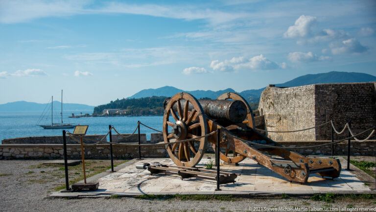 Large iron cannon on a heavy wooden carriage beside the sea, with sailboats and distant hills beyond the fortress wall.