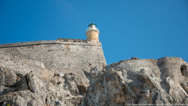 Cylindrical lighthouse with white gallery and green lantern roof above the rocky summit of Corfu's Old Fortress.