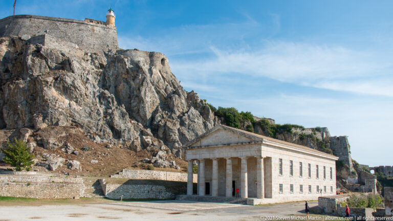 Church of Saint George beneath the steep rocky summit of the Old Fortress, with its Doric portico in the foreground.