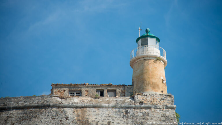 Close upward view of the Old Fortress lighthouse with ocher cylindrical tower, white gallery, green lantern roof, and adjoining masonry.