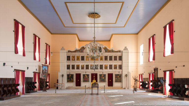 Centered nave of the Church of Saint George with chandelier, red-curtained windows, seating, and the 1864 stone iconostasis.