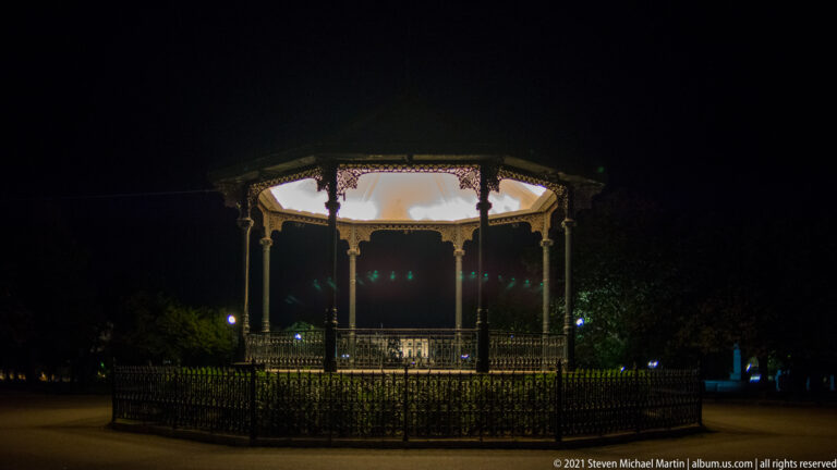Illuminated octagonal metal bandstand with decorative railings and roof, surrounded by a low fence in a dark park.