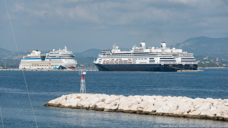 Two large cruise ships offshore beyond a white rock breakwater, with mountains rising in the background.