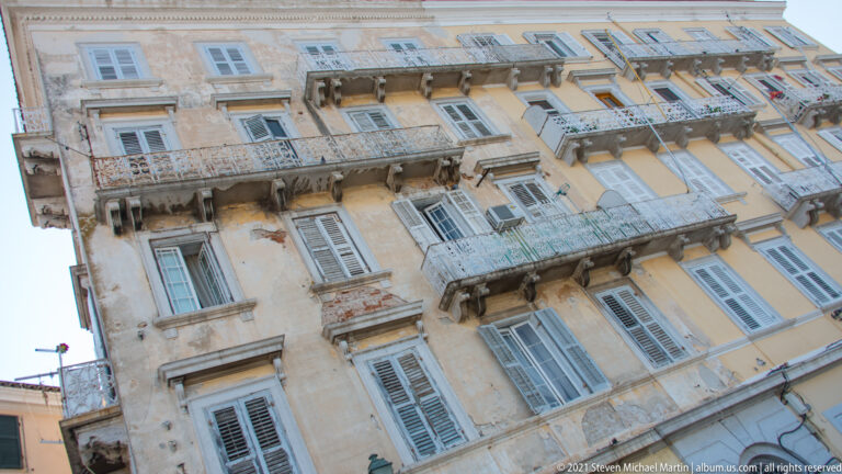 Upward view of a weathered ocher Corfu façade with many balconies, pale shutters, carved brackets, and air conditioners.