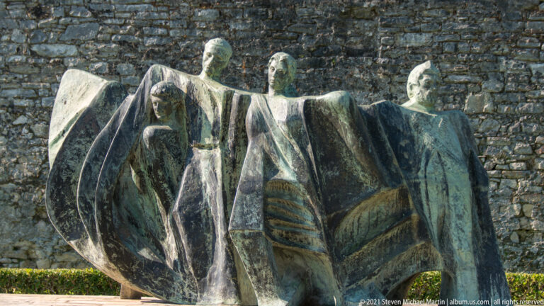 Bronze sculpture of four stylized human figures joined by broad cloak-like forms against a stone wall.