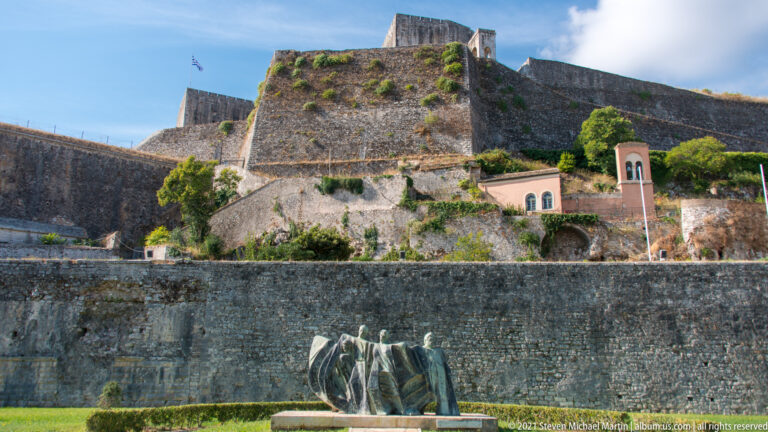Bronze group sculpture on a lawn below massive layered stone bastions and walls of Corfu's Old Fortress.