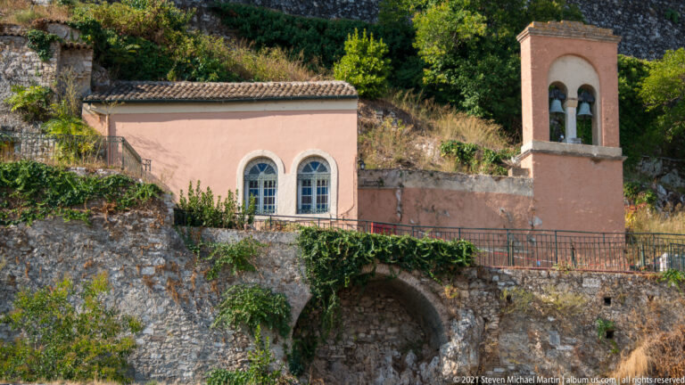 Small pink church-like building with paired arched windows and a two-bay bell tower set against fortress masonry.