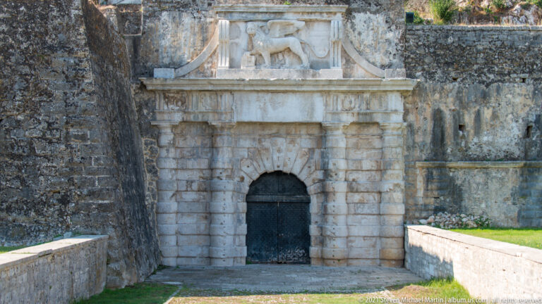 Ornate pale-stone fortress gateway with rusticated arch, black door, and sculpted pediment set in dark masonry.
