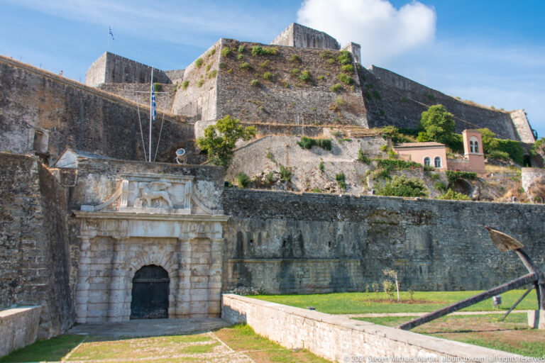 Ornate fortress portal below massive bastions, with a historic anchor on grass and a small pink bell tower at right.